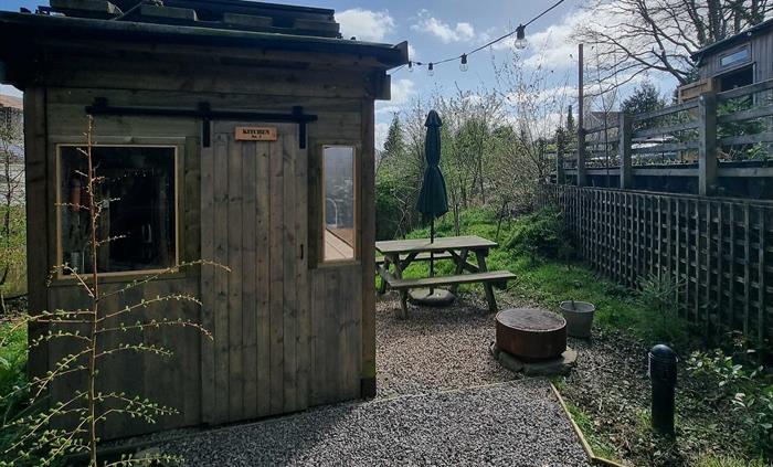 A cabin with a picnic table and firepit surrounded by greenery and a blue cloudy sky
